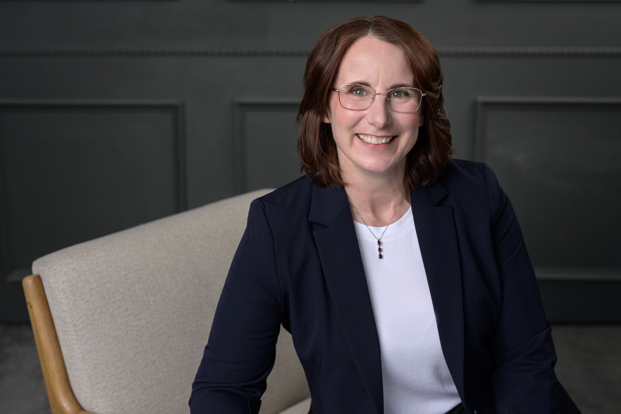 Smiling woman in a dark jacket and white top, seated on a beige chair against a gray wall, exuding professionalism and approachability.