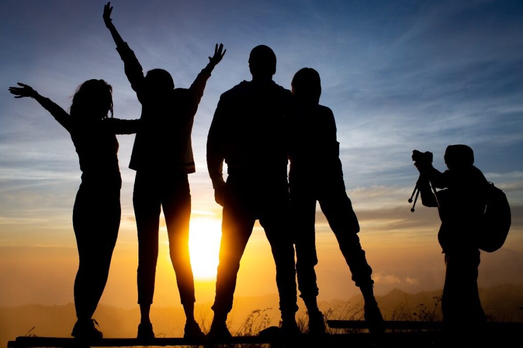 Silhouettes of five friends celebrating at sunset, with one person taking a photo. The scene captures a joyful moment in nature.