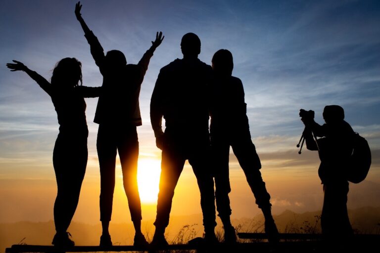 Silhouettes of five friends celebrating at sunset, with one person taking a photo. The scene captures a joyful moment in nature.