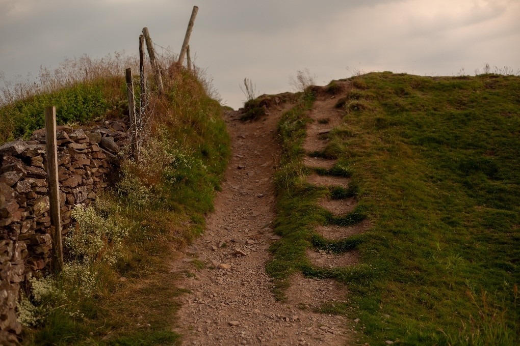 “A narrow dirt path winding uphill between a grassy slope and a rustic stone wall with wooden fence posts under a cloudy sky.