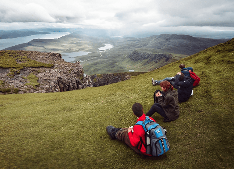 Hikers relax on a grassy hillside, overlooking a scenic landscape with mountains and a river in the background, highlighting nature's beauty.